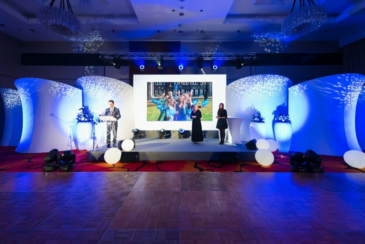 On a brightly lit stage with blue and white lighting is a large screen showing a group of people. There are three people on stage: one speaking at the podium, one standing to the right of the screen and one to the right. Photo coverage of the events highlights the large white balls decorating the stage.  