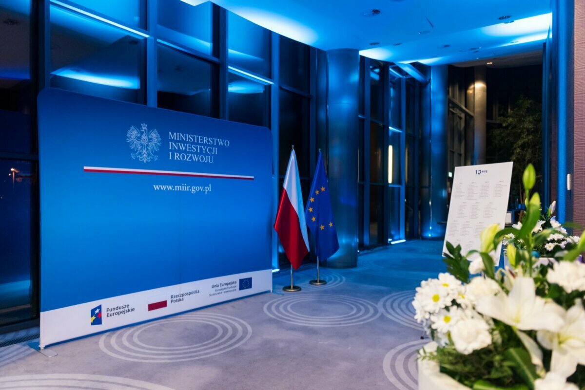 The blue background reads "Ministry of Investment and Development" along with the website URL "www.miir.gov.pl". In front are the flags of Poland and the European Union. Nearby a list stand, and to the right of the stacked white flowers is an event photo from Warsaw.  