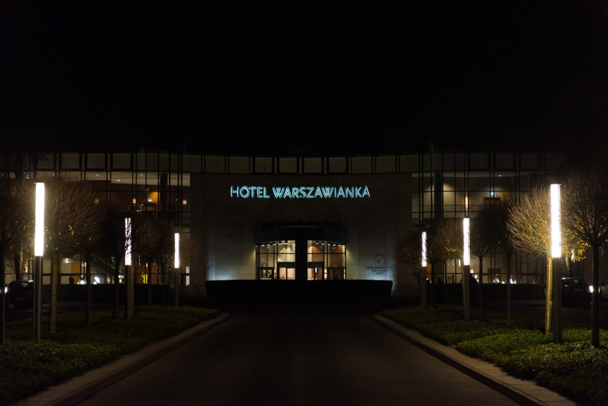 Night view of the front entrance to the Warszawianka Hotel, illuminated by tall lanterns. Above the entrance, the sign "HOTEL WARSZAWIANKA" is brightly illuminated in green. Captured by event photographer warszawa, this modern building with large glass windows and a well-kept driveway stands out beautifully.  