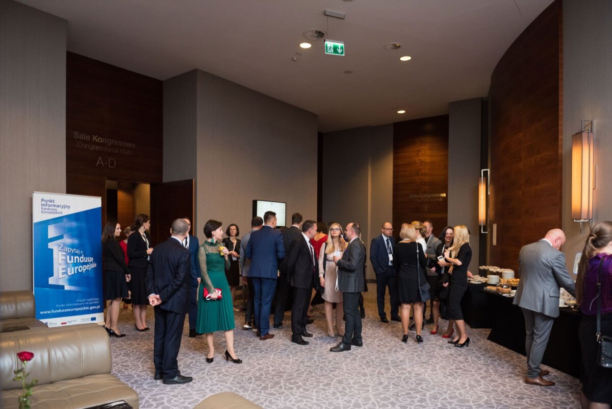 A group of people dressed in formal attire mingle in a spacious, modern room with beige and brown decor. Some are holding drinks and carrying on a conversation at a snack table. In the background, a large banner and several signs in the hallway are visible, perfect for photo coverage of events.  