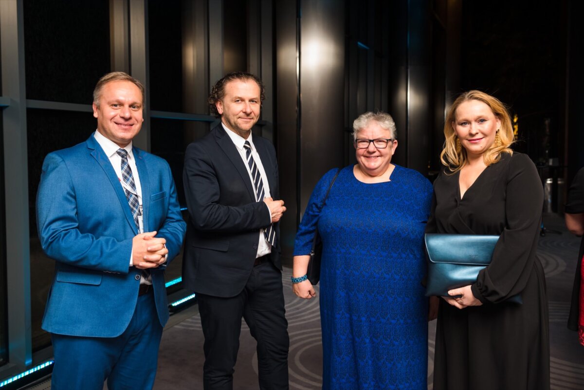 Four people are standing side by side in a formal setting, dressed in business attire. The person on the left is wearing a blue suit, next to him is a person in a black suit, then a person in a blue dress, and on the right is a person in a black dress. Perfect for event photography or if you are looking for an event photographer warsaw!  