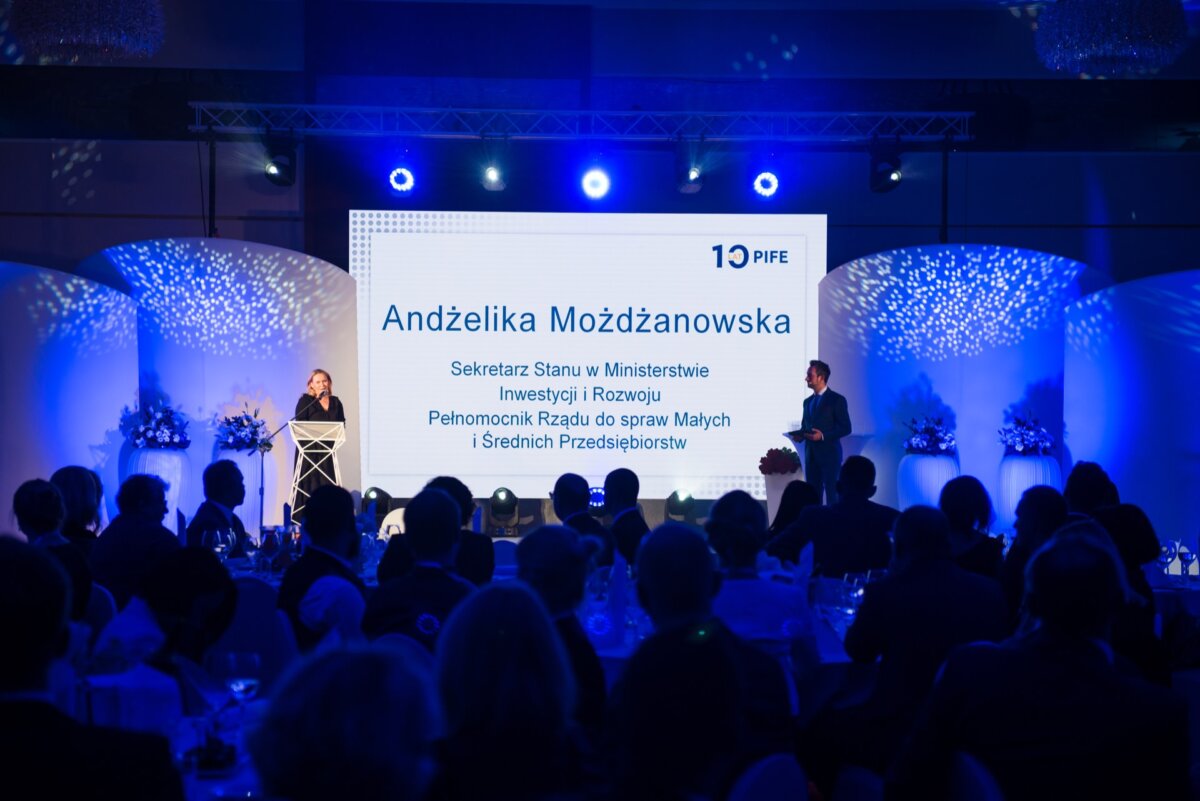 A woman stands on a stage at a well-lit conference or event and speaks into a microphone. Behind her, a large screen shows the name "Angelika Możdżanowska" and her title. The audience sits at the front, facing the stage, and an event photographer warszawa captures the moment. The setting is formal and professional.   