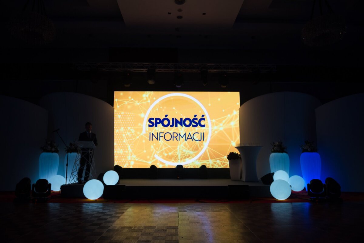 A dimly lit stage with a large LED screen displaying the inscription "INFORMATION SPOILER" on an abstract background. The stage, ideal for photo coverage of events, is decorated with illuminated white balls and modern furniture. To the left of the screen, a person stands on a podium.  