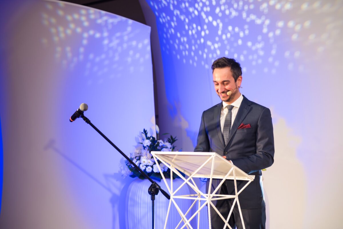 A man in a suit stands at a white podium and speaks into a microphone. He smiles, and a floral composition can be seen in the background. Blue and white lighting creates a patterned effect on the wall behind him, capturing the essence of event photography by an experienced event photographer Warsaw.  