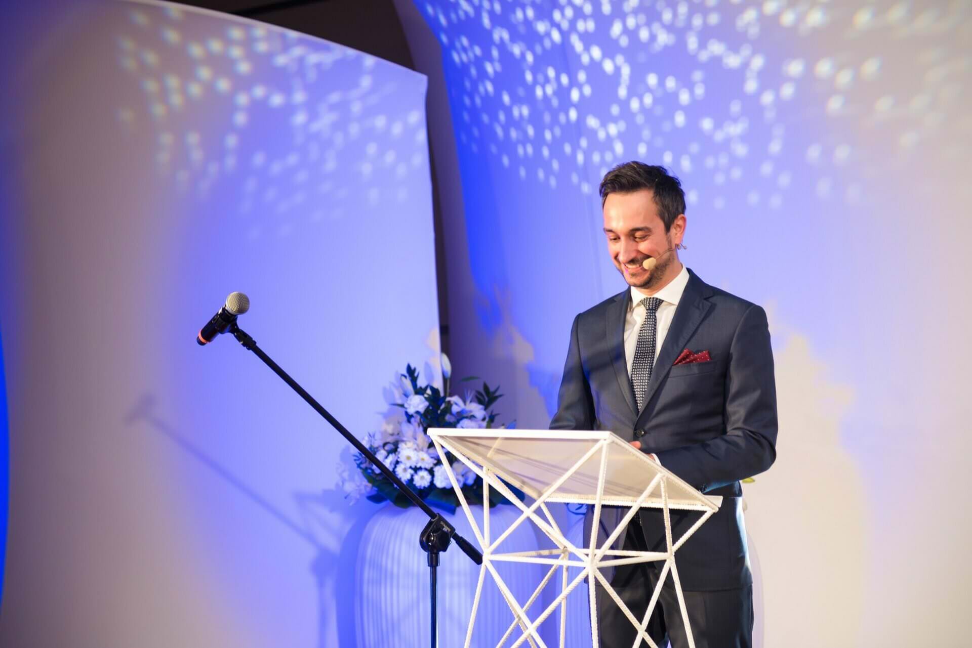 Ein Redner im Anzug spricht vom Podium auf der Galabühne, sein Gesicht ist trotz starker Bühnenbeleuchtung und blauem Hintergrund gut erkennbar.