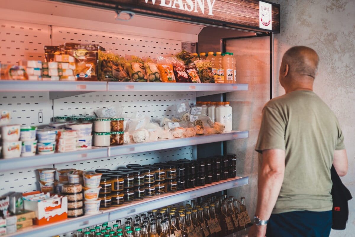 Łysy mężczyzna w zielonej koszulce przegląda półki wypełnione różnymi pakowanymi produktami spożywczymi i napojami w sklepie. Wśród przedmiotów znajdują się słoiki, butelki i różne pojemniki, starannie zorganizowane. Dobrze oświetlone półki eksponują asortyment dostępnych produktów, jakby uchwycony w oprawie fotografii eventowej.