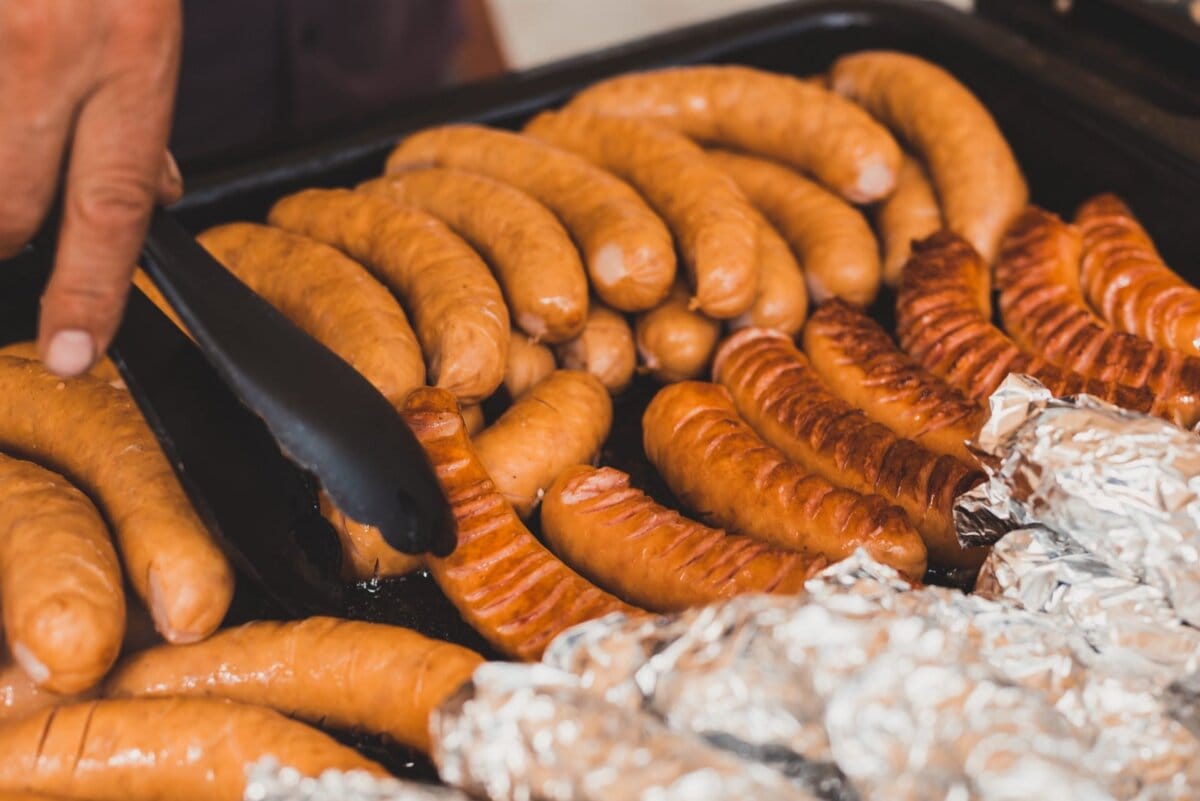 Ręką za pomocą szczypiec ułóż złotobrązowe i lekko zwęglone kiełbaski na grillu, obok kilku owiniętych w folię produktów. Ten moment oddaje istotę fotografii eventowej, idealnej do tętniącej życiem fotorelacji z wydarzeń w sercu Warszawy.