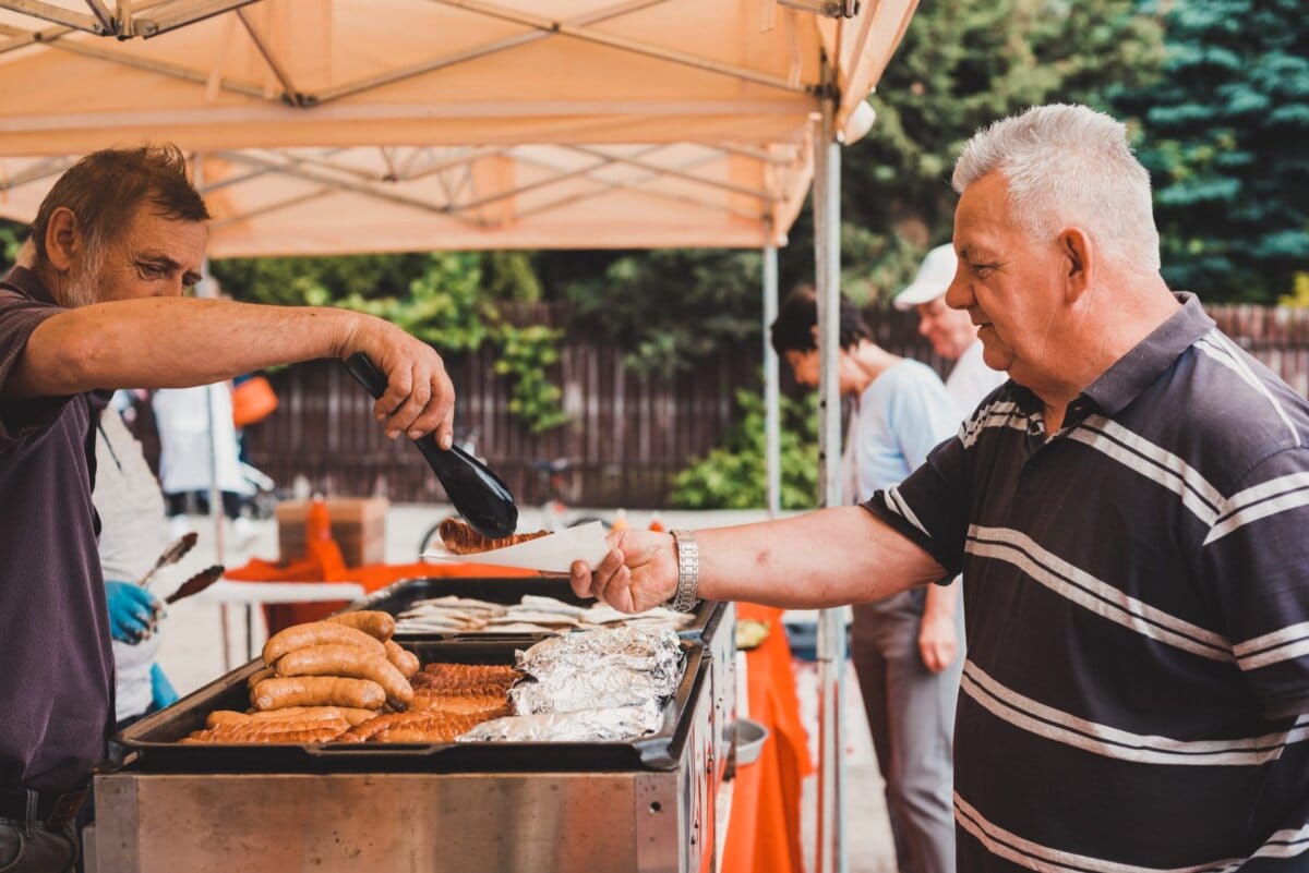 Mężczyzna w pasiastej koszuli trzyma talerz, podczas gdy inny mężczyzna w ciemnej koszuli podaje kiełbaski ze stoiska z jedzeniem pod namiotem. Na straganie można znaleźć kiełbaski i produkty przykryte folią. Na tej fotografii eventowej ludzie stoją w tle na świeżym powietrzu, z widocznymi drzewami i płotem, oddając ożywioną atmosferę.