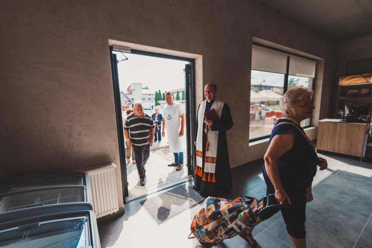 Mężczyzna w stroju religijnym stoi w pobliżu otwartych drzwi budynku, prawdopodobnie kościoła lub domu kultury. Wchodzi kilka osób, w tym kobieta z kraciastą torbą na zakupy na kółkach, inne zbierają się na zewnątrz. Atmosfera wydaje się swobodna i przyjazna, idealna na fotografię eventową.