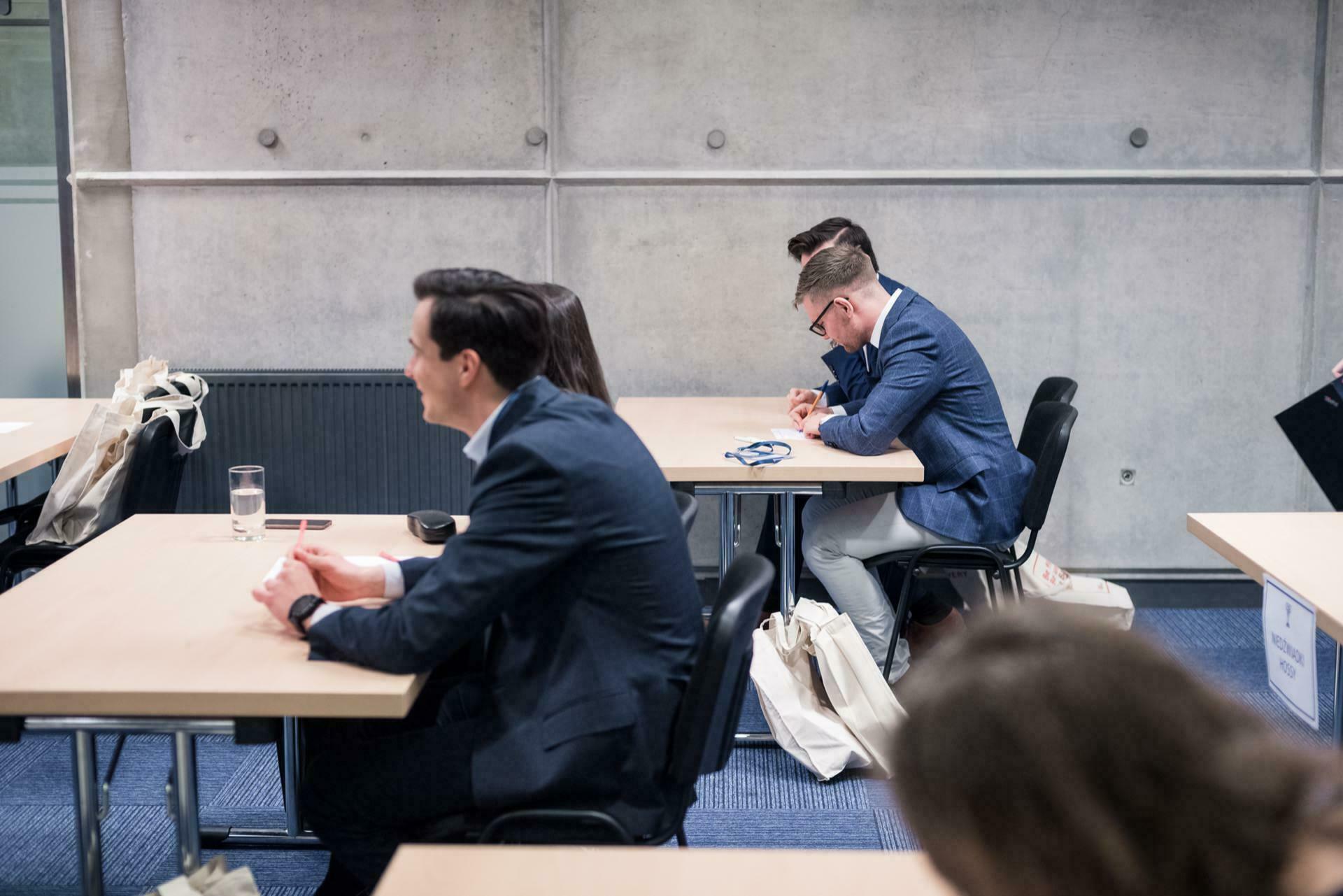 People are sitting at tables in a classroom or conference room. Some are busy talking, others are focused on the papers lying in front of them. The room has a concrete wall and blue carpet. The event participants dressed in business attire create the perfect scene for event photography.   