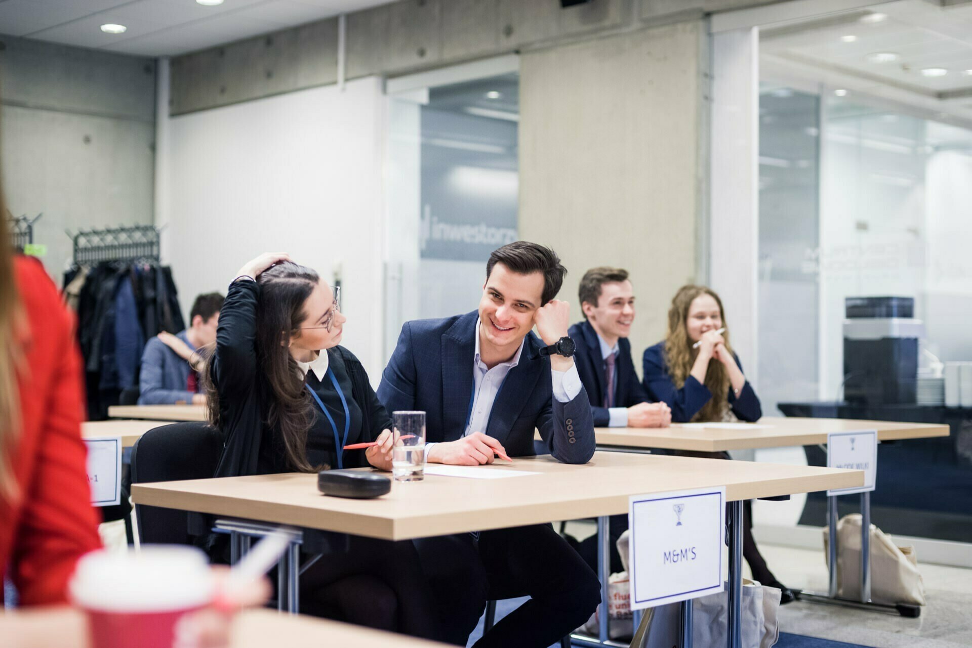 A group of young professionals sit at desks in a modern office space, talking and smiling. One man in the foreground interacts with the woman next to him, and both seem cheerful. The atmosphere is casual and collaborative - the perfect scene for a photo event.  