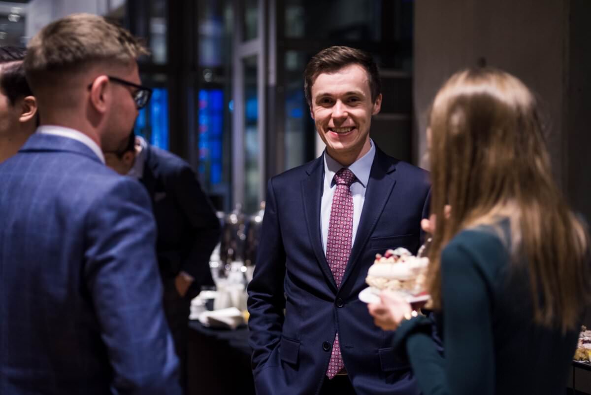 Eine Gruppe von formell gekleideten Personen unterhält sich bei einer gesellschaftlichen Veranstaltung. Einer der Männer, der einen dunklen Anzug und eine rote Krawatte trägt, lächelt direkt in die Kamera und fängt damit die Essenz des Eventfotografen Warschau ein. Andere Personen um ihn herum unterhalten sich angeregt und eine Frau, die einen Kuchenteller hält, ist teilweise im Hintergrund zu sehen.
