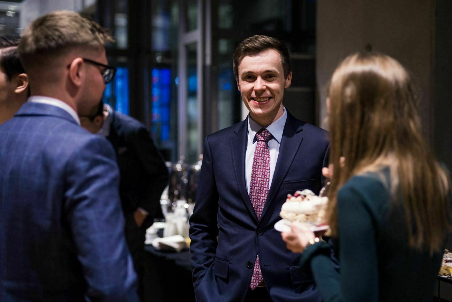 A group of people in formal attire are chatting at a social event. One of the men, dressed in a dark suit and red tie, smiles directly at the camera, capturing the essence of event photographer Warsaw. Other people around him are busy chatting, and a woman holding a plate of cake is partially visible in the back.  