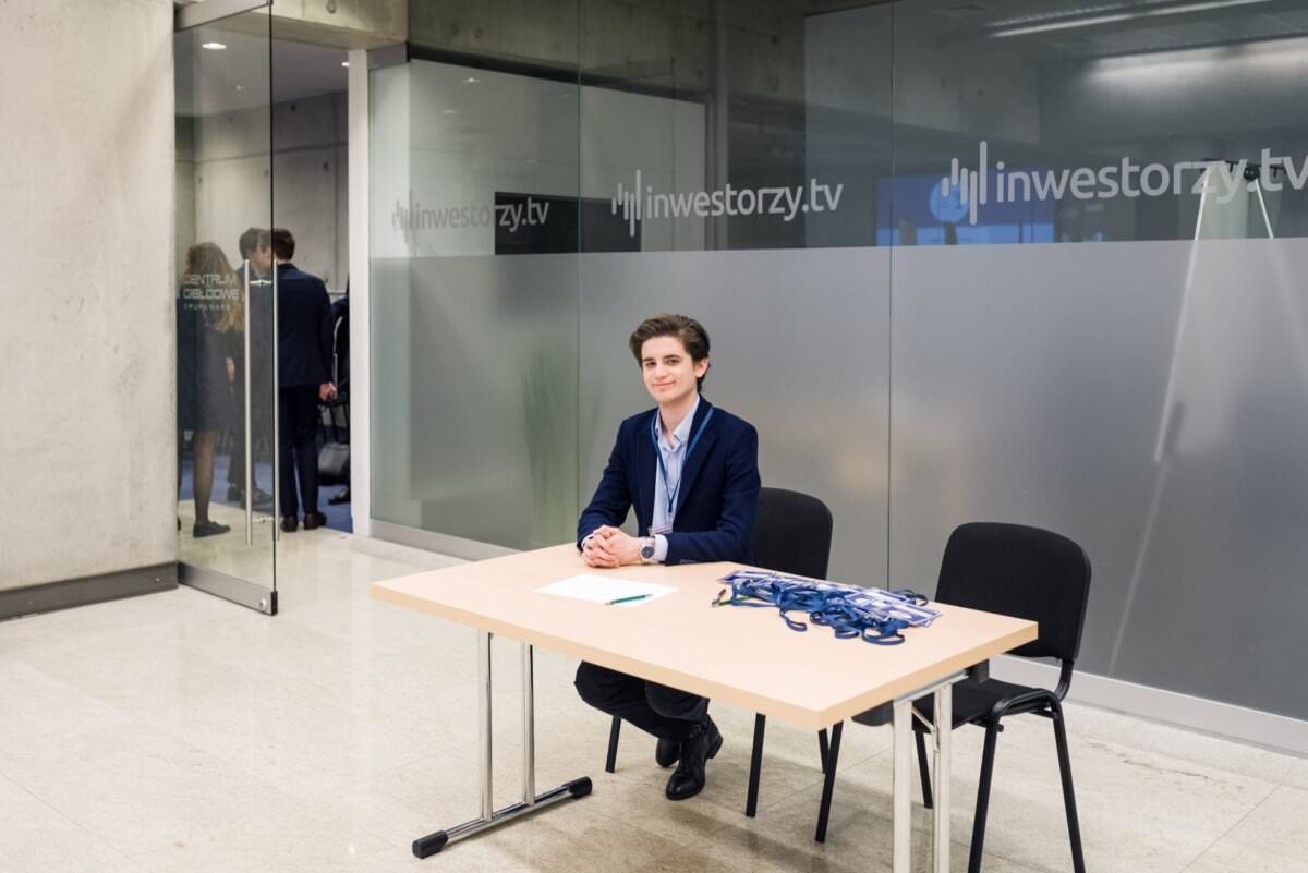Eine Person im Geschäftsanzug sitzt hinter einem Tisch mit Namensschildern an der Konferenzregistrierung. Der Raum hat Glaswände, die mit dem Logo von 'inwestorzy.tv' verziert sind. Im Hintergrund, durch die Tür hindurch, sind weitere Personen zu sehen, die Teil der Fotoreportage über die Veranstaltungen sind.