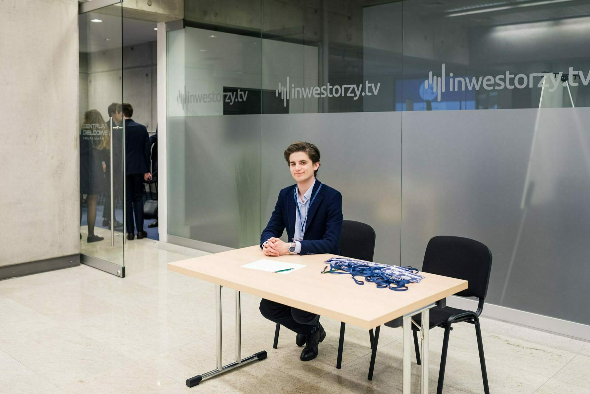 A person in business attire sits behind a table with badges at the conference registration desk. The room has glass walls decorated with the "investors.tv" logo. In the background, through the door, other people can be seen as part of the photo coverage of the events.  