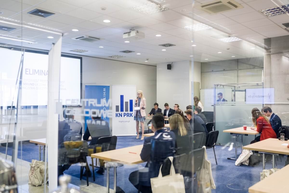 Menschen nehmen an einer Fachkonferenz teil, die in einem modernen Büro stattfindet. Die Präsentationen werden auf Bildschirme projiziert, während die Teilnehmer an Tischen sitzen und sich mit dem Material beschäftigen. Der Redner an der Stirnseite spricht zum Publikum und das Branding des Unternehmens ist sichtbar, alles eingefangen von einem Eventfotografen Warschau.
