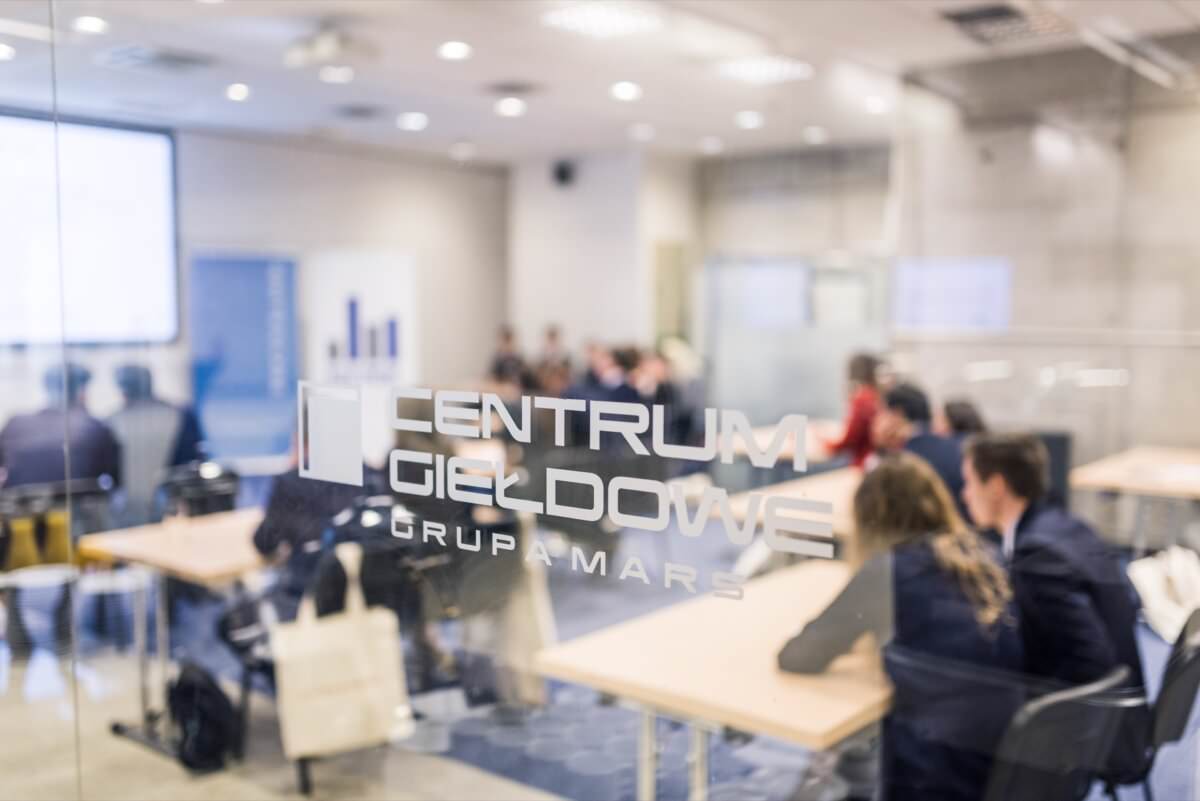 Ein Konferenzraum, in dem mehrere Personen an Tischen sitzen und diskutieren oder aufmerksam zuhören. Die Bühne ist durch eine Glastür zu sehen, die die Aufschrift "CENTRUM GIED GRUPA MAR S." trägt. Im Hintergrund ist ein Präsentationsbildschirm zu sehen, der die Essenz der Veranstaltung einfängt Fotoreportage von Eventfotograf warszawa.