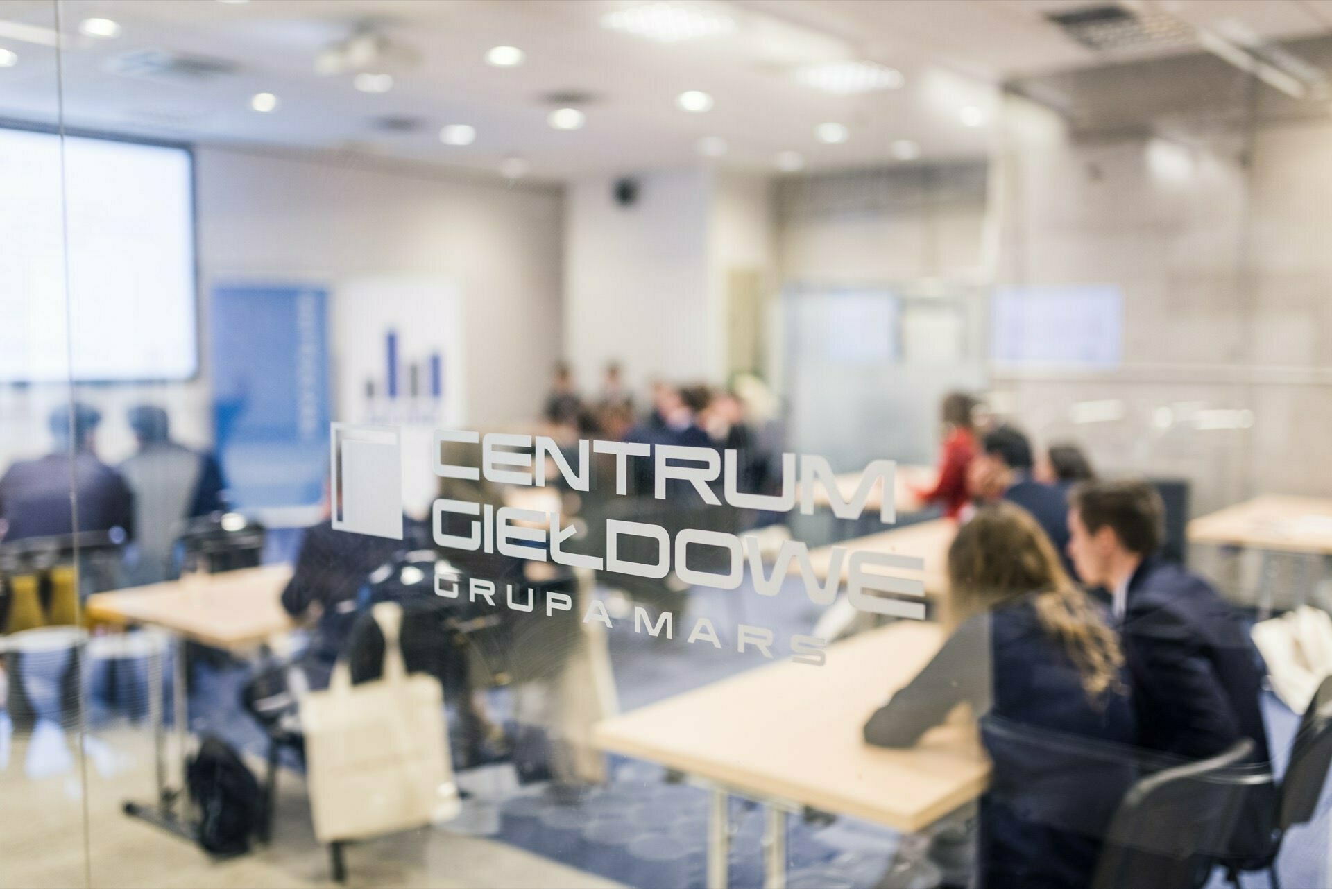 A conference room with several people sitting at tables discussing or listening attentively. The scene is viewed through a glass door, which bears the inscription "MAR S. GROUP GIEZ CENTER." A presentation screen is visible in the background, capturing the essence of the event photo report by event photographer warszawa.