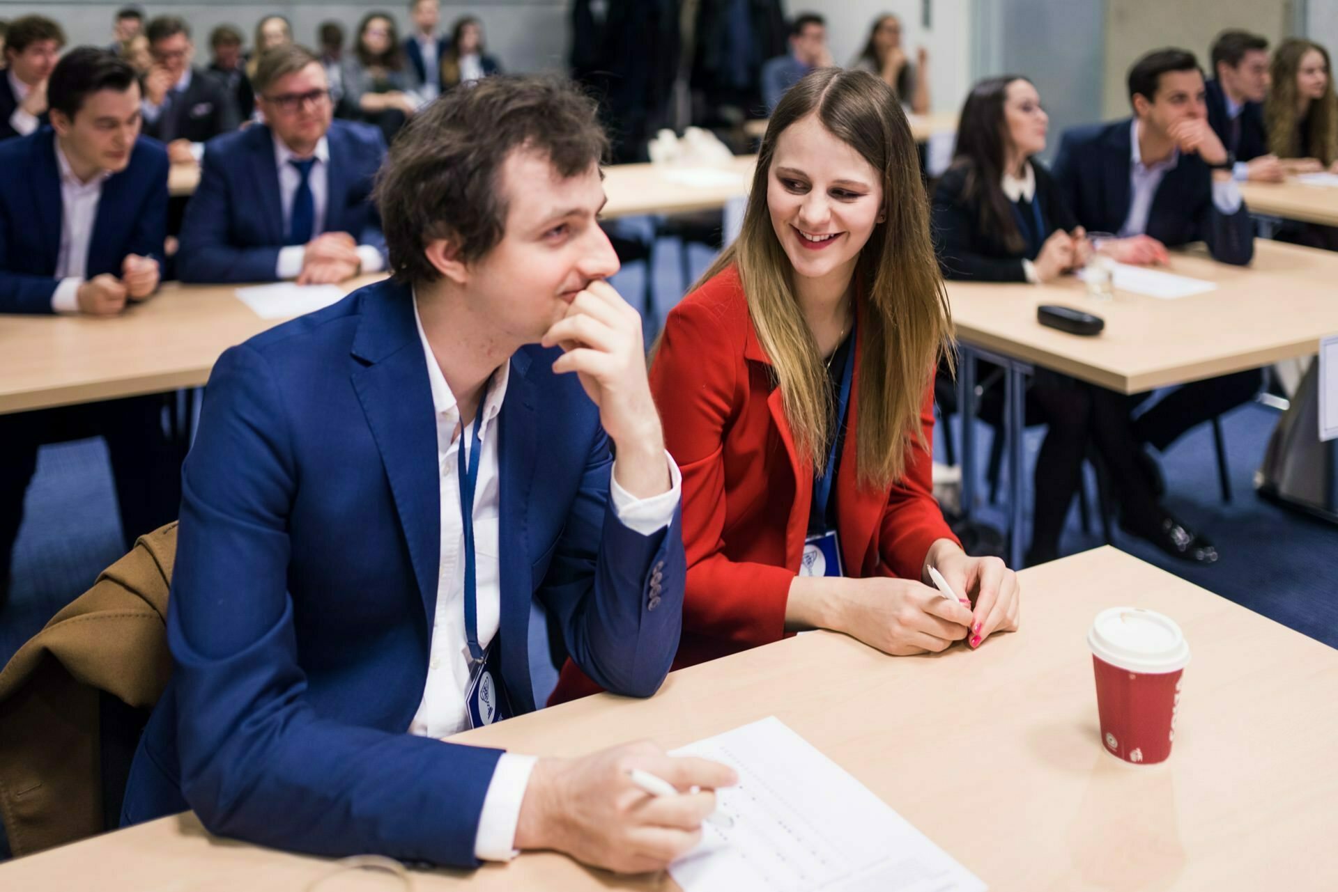 A man in a blue suit and a woman in a red jacket sit at a table in a conference room and engage in conversation. The room, captured as part of the event photo essay, is filled with other event participants sitting at desks, with papers and a cup of coffee visible on the man's table. 