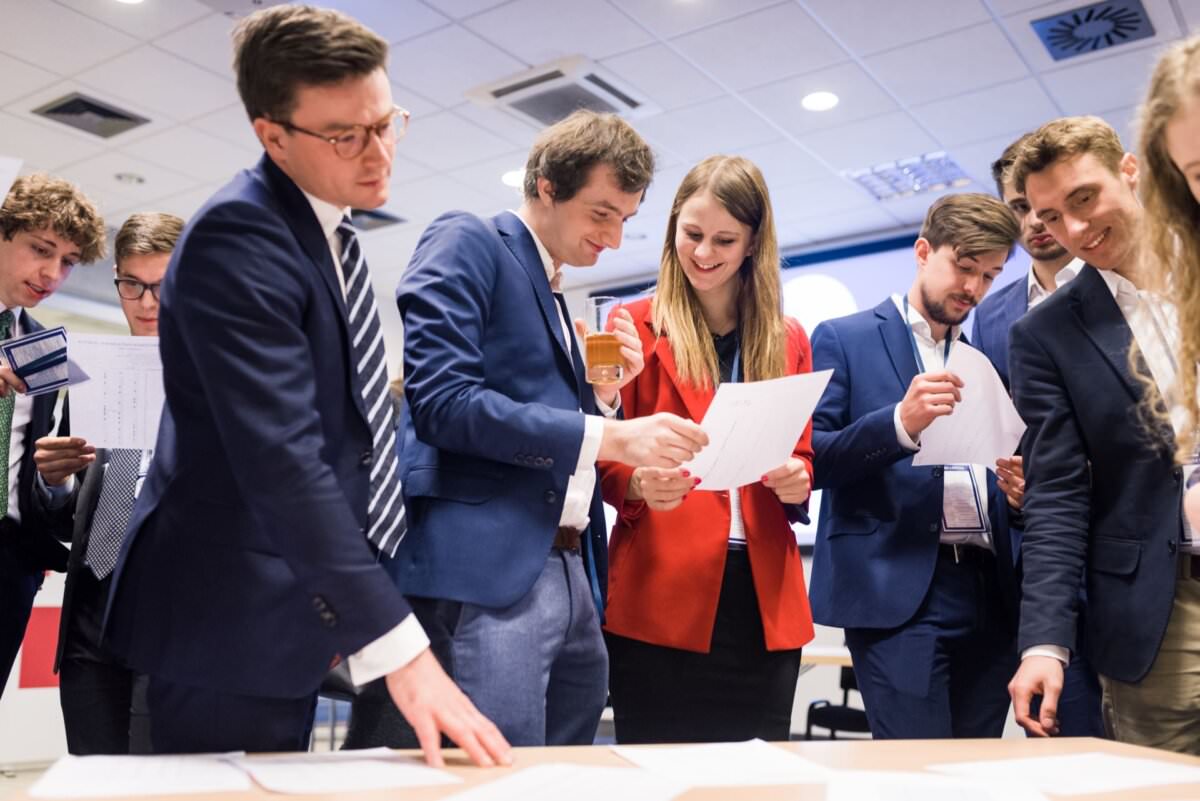 Eine Gruppe von Menschen in Geschäftskleidung versammelt sich um einen Tisch und prüft verschiedene Dokumente. Einige halten Dokumente in der Hand, andere sind in ein Gespräch vertieft. Die Umgebung sieht sehr geschäftlich aus, mit Deckenleuchten und Lüftungsschächten über dem Kopf, die den perfekten Moment für die Eventfotografie von Eventfotograf Warszawa einfangen.