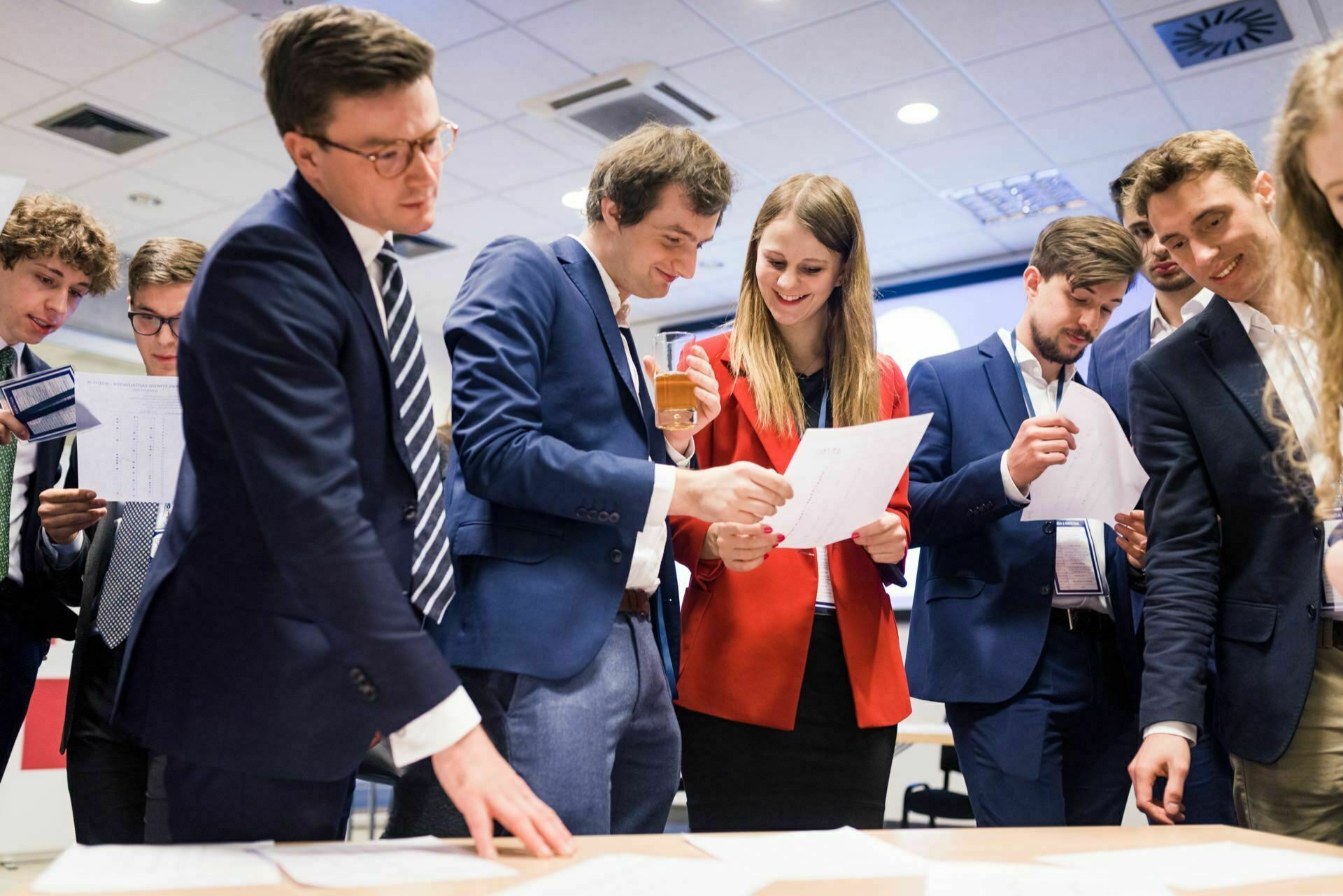 A group of people in business attire gather around a table, reviewing various documents. Some are holding documents, others are engaged in conversation. The environment looks corporate, with ceiling lights and vents visible overhead, capturing the perfect moment for event photography by event photographer warsaw.  