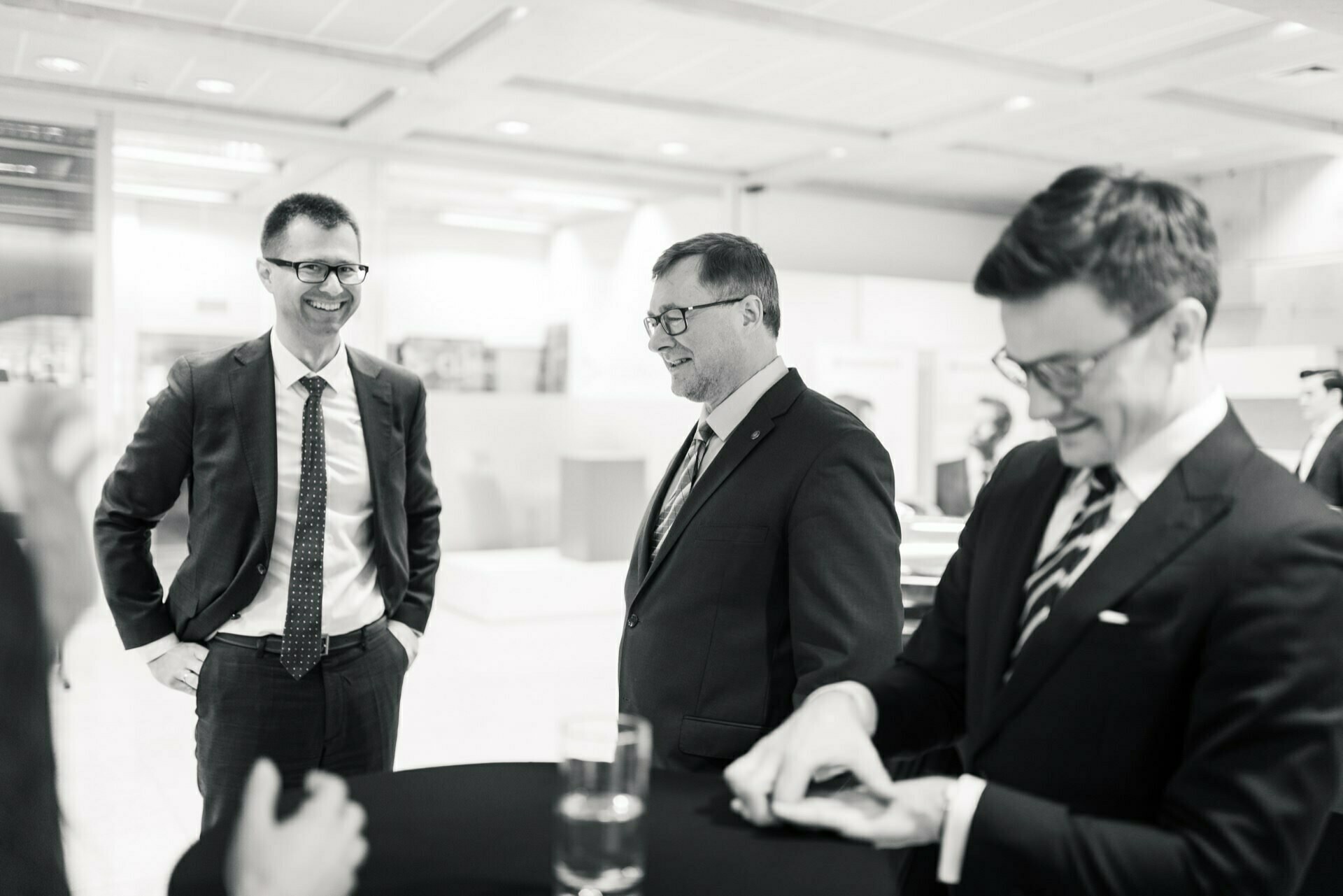 The black-and-white photo shows a social gathering: three men in suits around a table. One man is laughing, another is looking at his hands, and the third is smiling while keeping his hands in his pockets. This event photo shows the timeless elegance that only event photography can provide.  