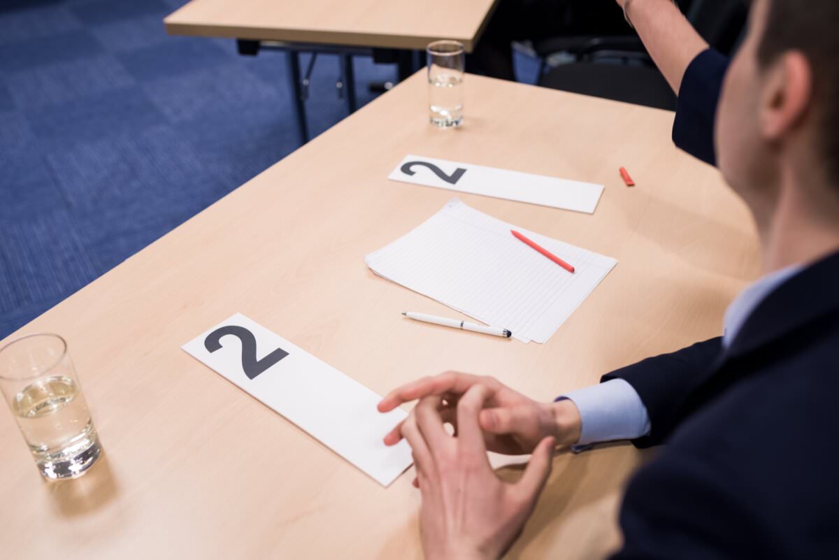 Eine Person sitzt an einem Schreibtisch mit nummerierten Karten mit der Aufschrift '2', einem Blatt liniertem Papier, einem roten und einem schwarzen Stift. Zwei Wassergläser stehen auf dem Schreibtisch. Ein weiterer Schreibtisch mit ähnlichen Gegenständen ist teilweise im Hintergrund zu sehen; es sieht aus, als ob die Vorbereitungen für ein Veranstaltungsfoto im Gange sind.