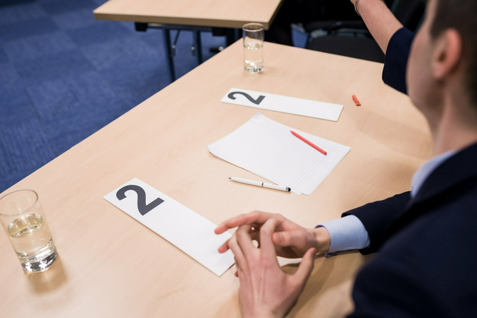 A person sits at a desk with numbered cards marked "2", a sheet of lined paper, a red and black pen. Two glasses of water stand on the desk. Another desk with similar items is partially visible in the background; it looks like preparations for an event photograph are underway.  