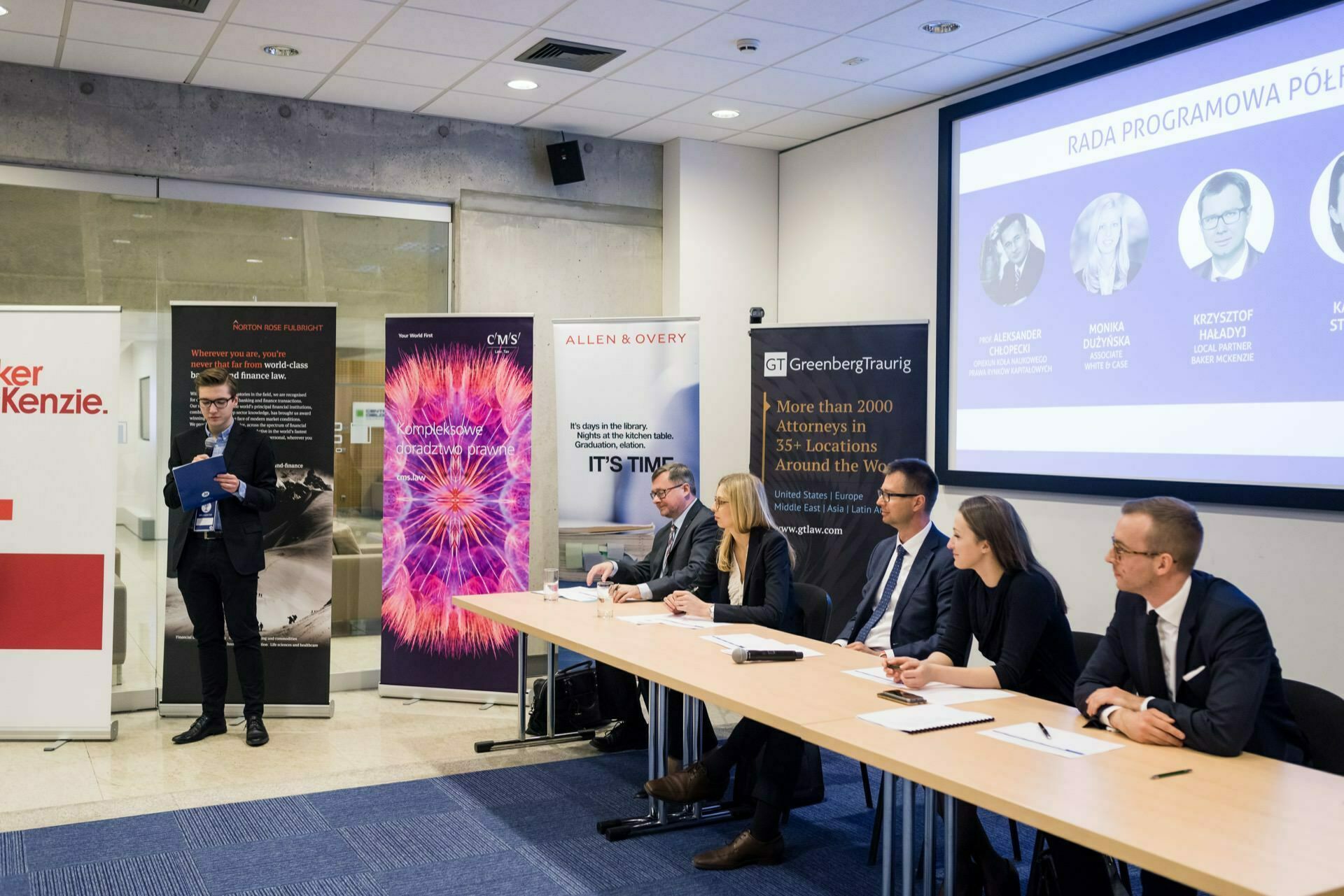 The speaker is addressing a panel seated at a long table in a conference room. Behind the panelists are banners and a large screen displaying the list. The panelists are attentive, with notepads and microphones in front of them recording every detail of the photo-report of events.  