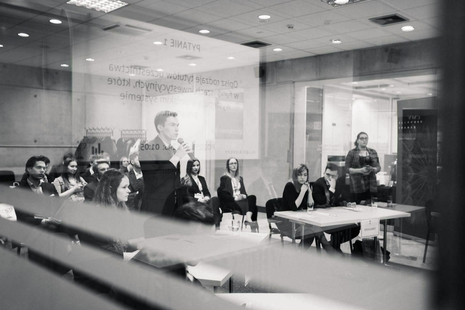 The black-and-white photo shows a group of people sitting in a room, seen through a glass with reflections. One person is standing and speaking into a microphone. Several people appear to be engaged or taking notes, illustrating a fascinating photo report of events by an event photographer warsaw.  