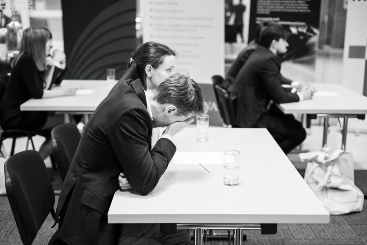 Das Schwarz-Weiß-Foto zeigt Menschen, die an Tischen sitzen und in schriftliche Arbeit vertieft sind. Der Mann im Vordergrund stützt seinen Kopf auf seine Hand und erweckt den Eindruck, gestresst oder müde zu sein. Auch andere Personen sind im Fokus und schaffen eine Studioatmosphäre in einer professionellen Umgebung - eine perfekte Fotoreportage der Ereignisse.