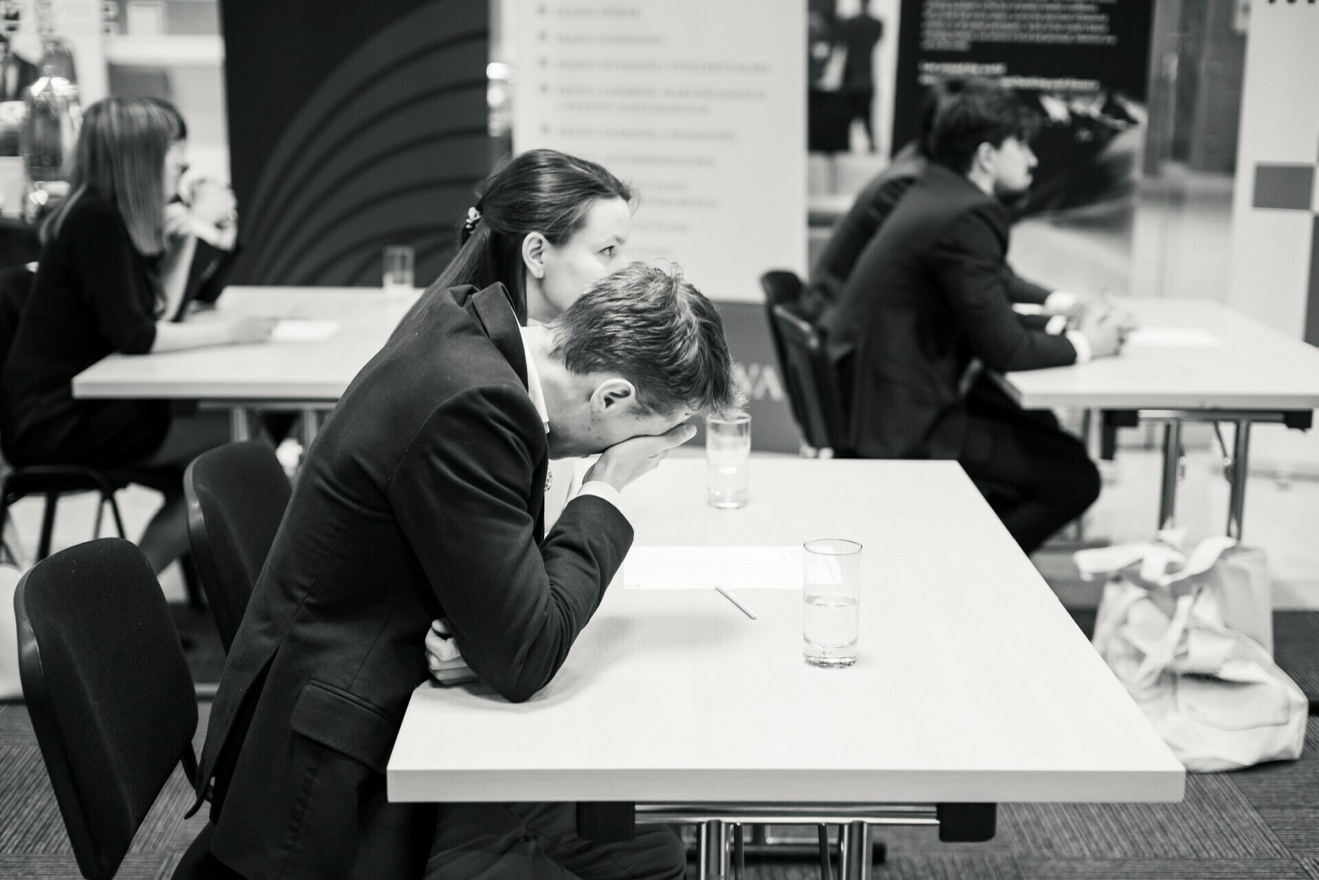 The black and white photo shows people sitting at tables, immersed in written work. The man in the foreground rests his head on his hand and gives the impression of being stressed or tired. Other people are also in focus, creating a studio atmosphere in a professional setting - an excellent photo report of the events.  