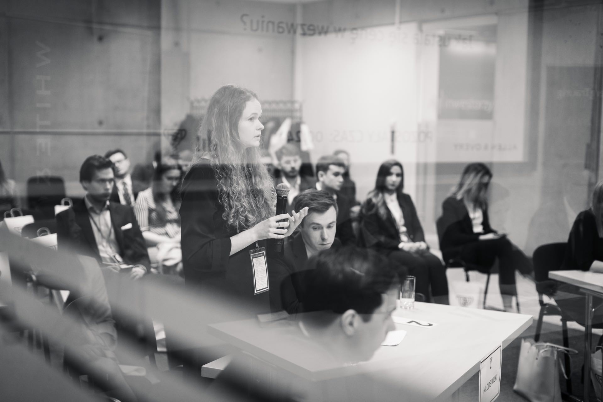 The black and white image of the conference room shows a woman speaking into a microphone. She faces a group of seated attendees, most of whom seem to be listening intently. Viewed through glass with a slightly blurred effect, this is an example of event photography in Warsaw.  