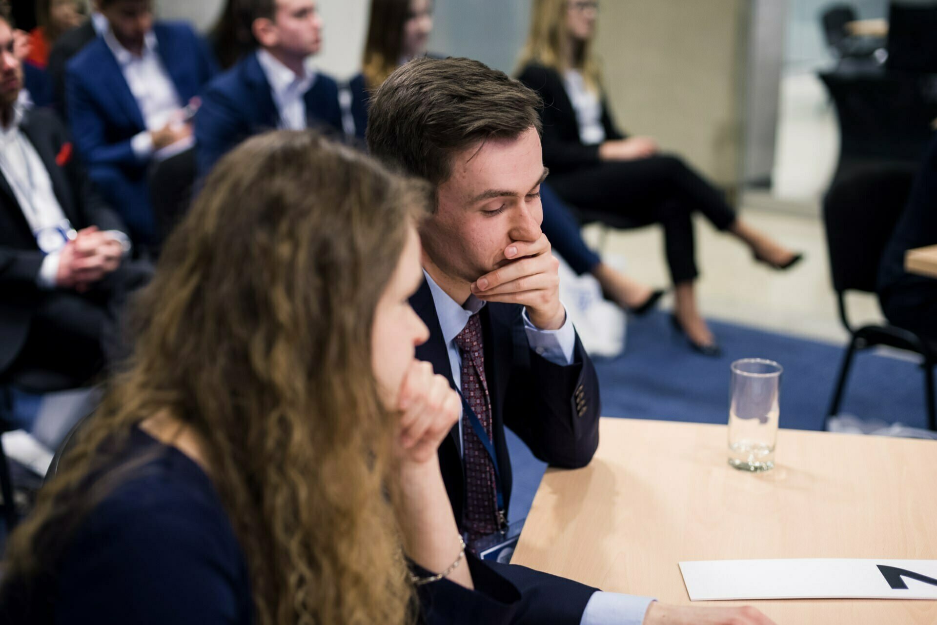 Sitting in a conference room is a group of people dressed in formal business attire. We focus on two people in the foreground, looking deeply thoughtful, with their hands resting on their chins, while others in the background listen intently to a speaker from outside the frame. This photo recap of the events perfectly captures the intensity and engagement of the moment.  