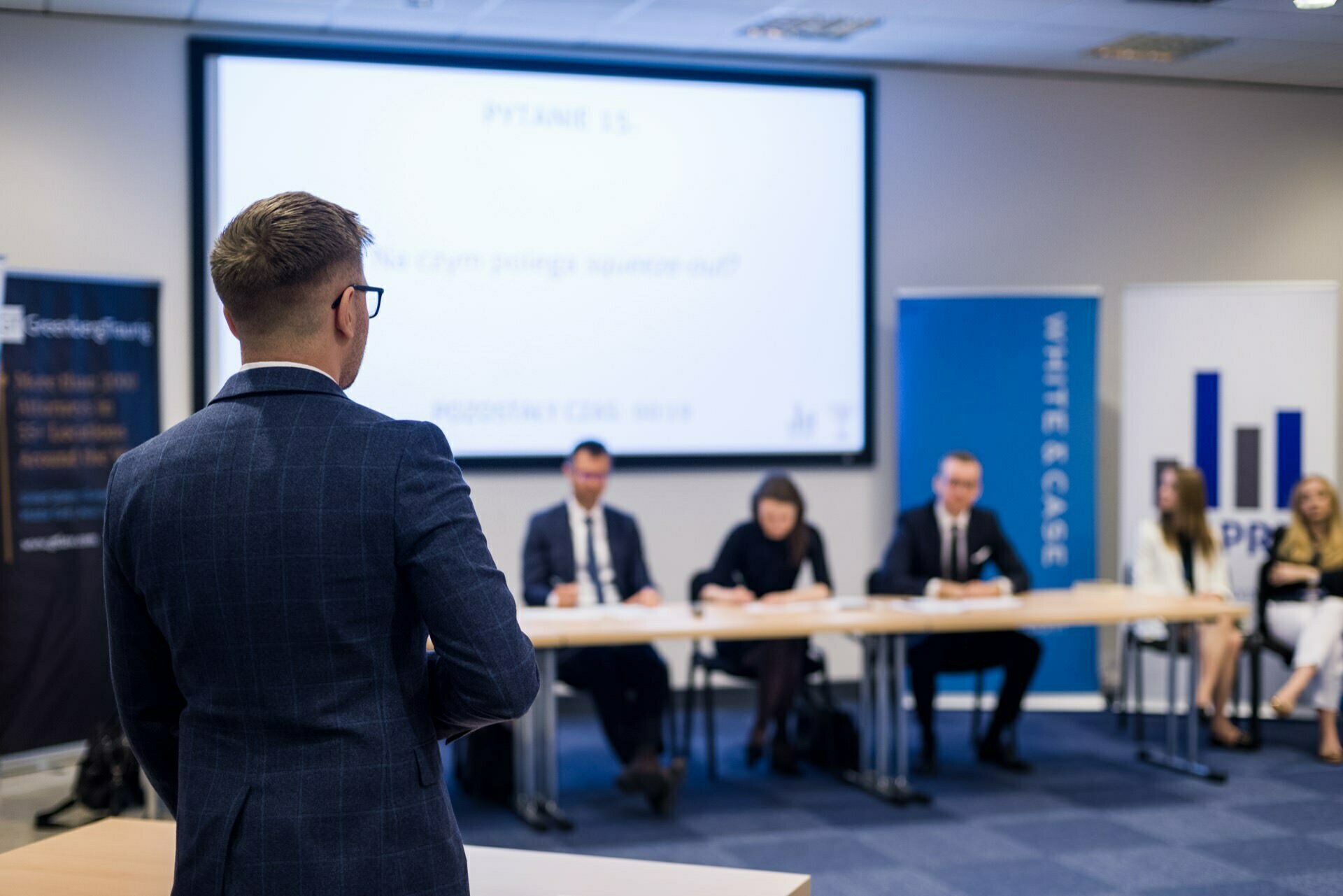 A man in a blue suit is facing a panel of people seated at a table in a brightly lit conference room. Behind the panel is a projection screen displaying text, and logo banners are visible on either side of the room. This photo report of the events perfectly captures the professionalism and atmosphere.  