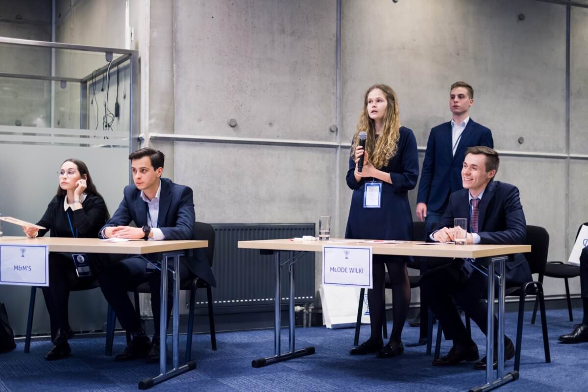 An der Debatte nimmt eine Gruppe von vier professionell gekleideten jungen Erwachsenen teil. Zwei Männer und eine Frau sitzen an Tischen, eine Frau steht und spricht energisch. Das moderne, industrielle Design des Raums mit seinen Betonwänden ist perfekt für jeden Eventfotografen oder Eventfotograf warszawa, der dynamische Momente einfangen möchte.