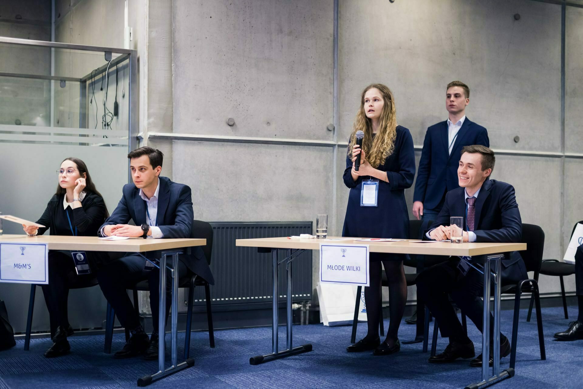 The debate involves a group of four professionally dressed young adults. Two men and a woman sit at tables, one woman stands and speaks energetically. The modern, industrial design of the room with concrete walls is perfect for any event photographer or event photographer warsaw who wants to capture dynamic moments.  