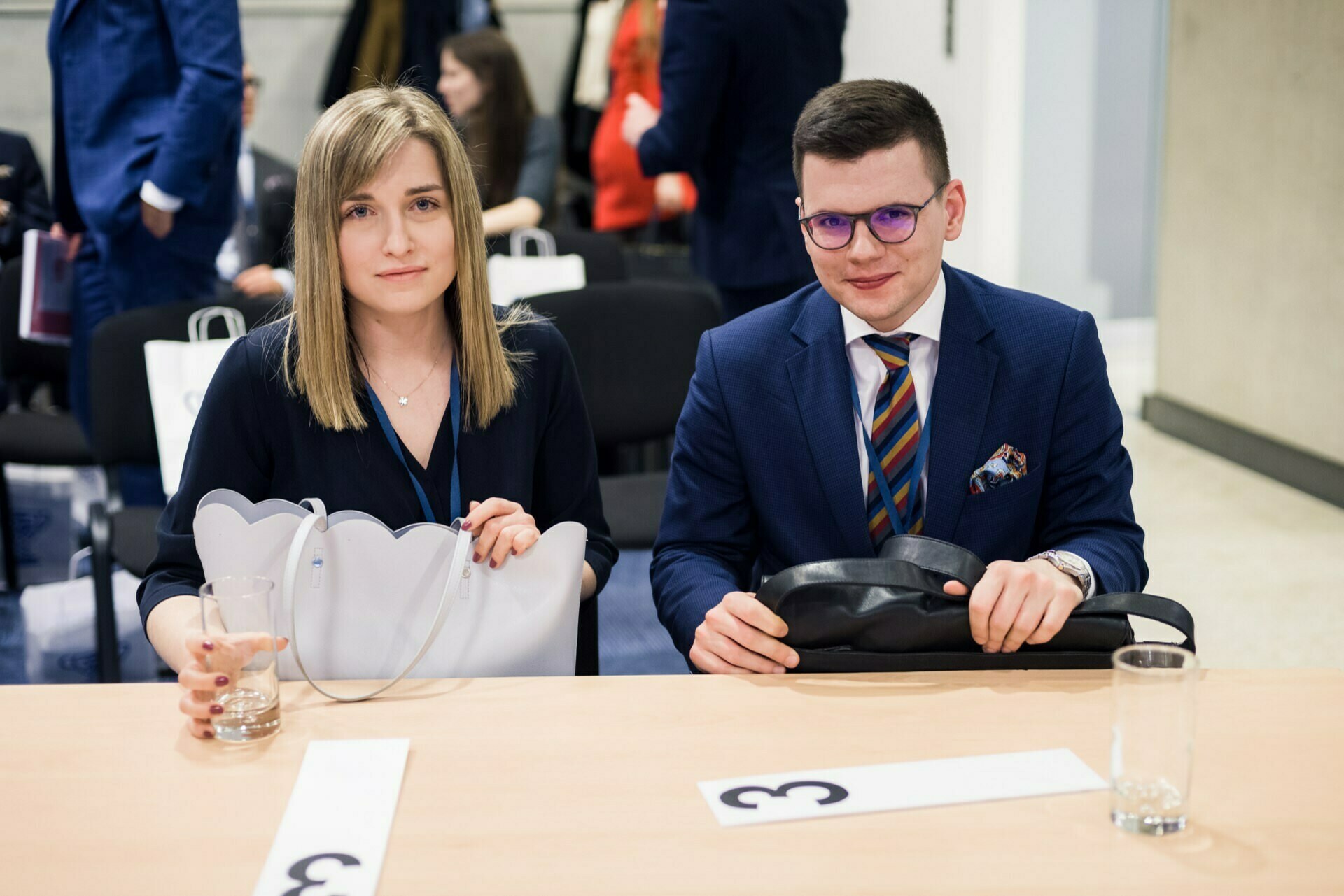 Sitting at a conference table are a woman and a man in formal attire, both holding black bags. The woman has a glass of water in front of her. They are smiling at the camera, and other participants can be seen in the background, capturing the perfect moment for event photography.  