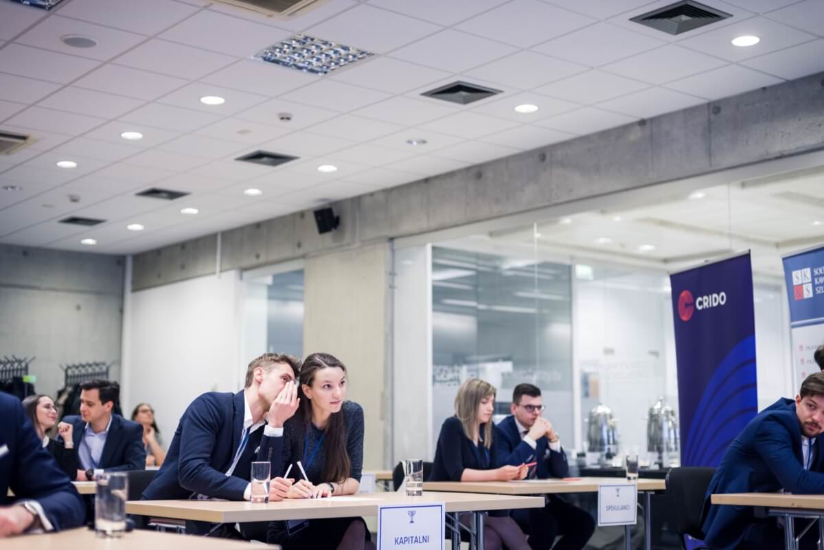 Eine Gruppe von Menschen in Geschäftskleidung sitzt an Tischen in einem modernen, gut beleuchteten Konferenzraum. Einige sind mit Gesprächen beschäftigt, andere scheinen sich auf ihre Aufgaben zu konzentrieren. Im Hintergrund sind Banner und gläserne Büros zu sehen, perfekt eingefangen vom Eventfotografen Warschau.