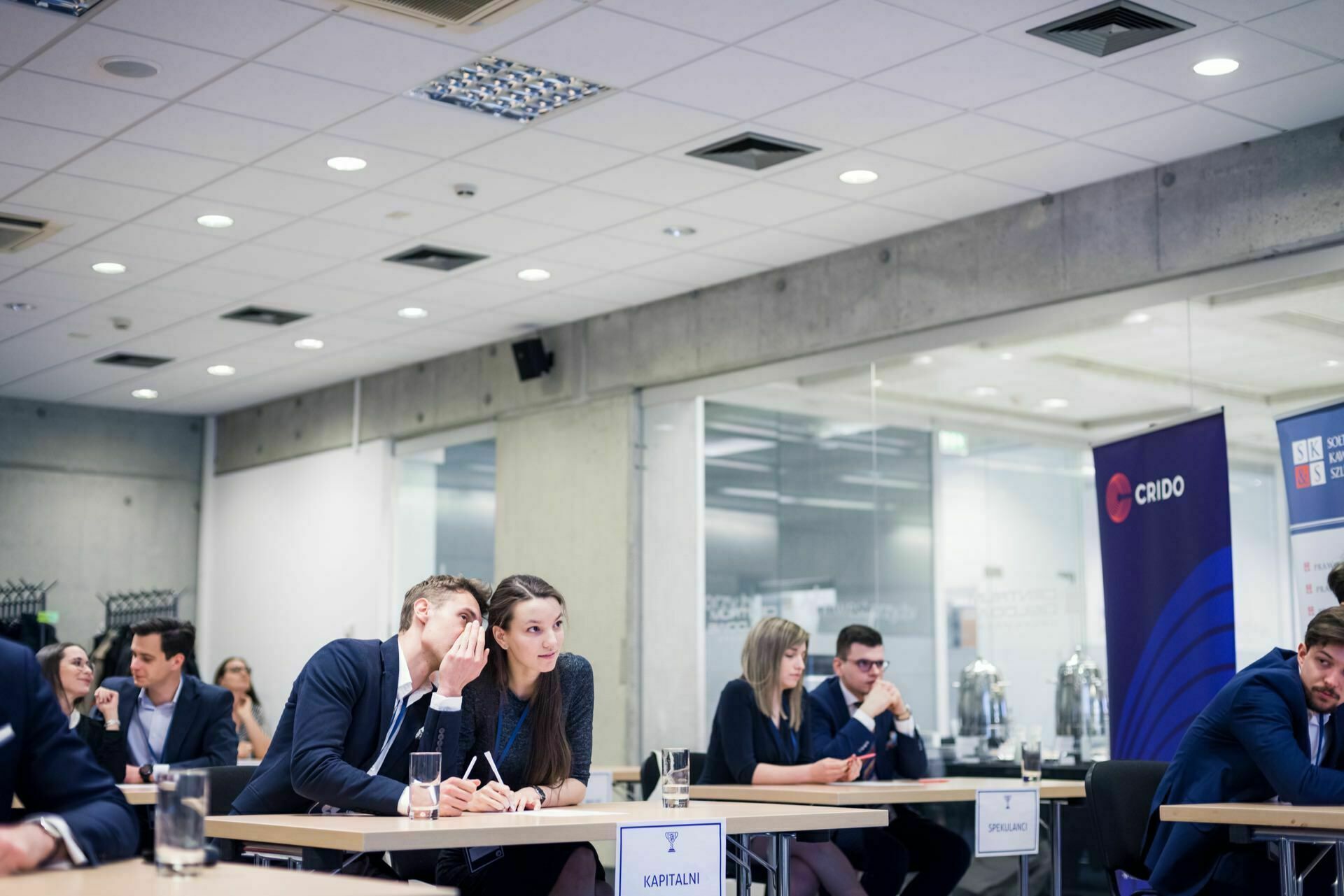 A group of people dressed in business attire sitting at tables in a modern, well-lit conference room. Some are busy talking, others seem focused on their tasks. Banners and glass offices are visible in the background, perfectly captured by event photographer Warsaw.  