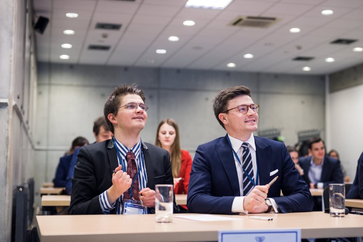 Eine Gruppe von Menschen in Geschäftskleidung sitzt an Schreibtischen in einem Konferenzraum. Zwei Männer im Vordergrund lächeln und sehen engagiert aus, einer ballt enthusiastisch die Fäuste. Die moderne, professionelle Umgebung unterstreicht die Fotoreportage der Ereignisse mit grauen Wand- und Deckenleuchten.