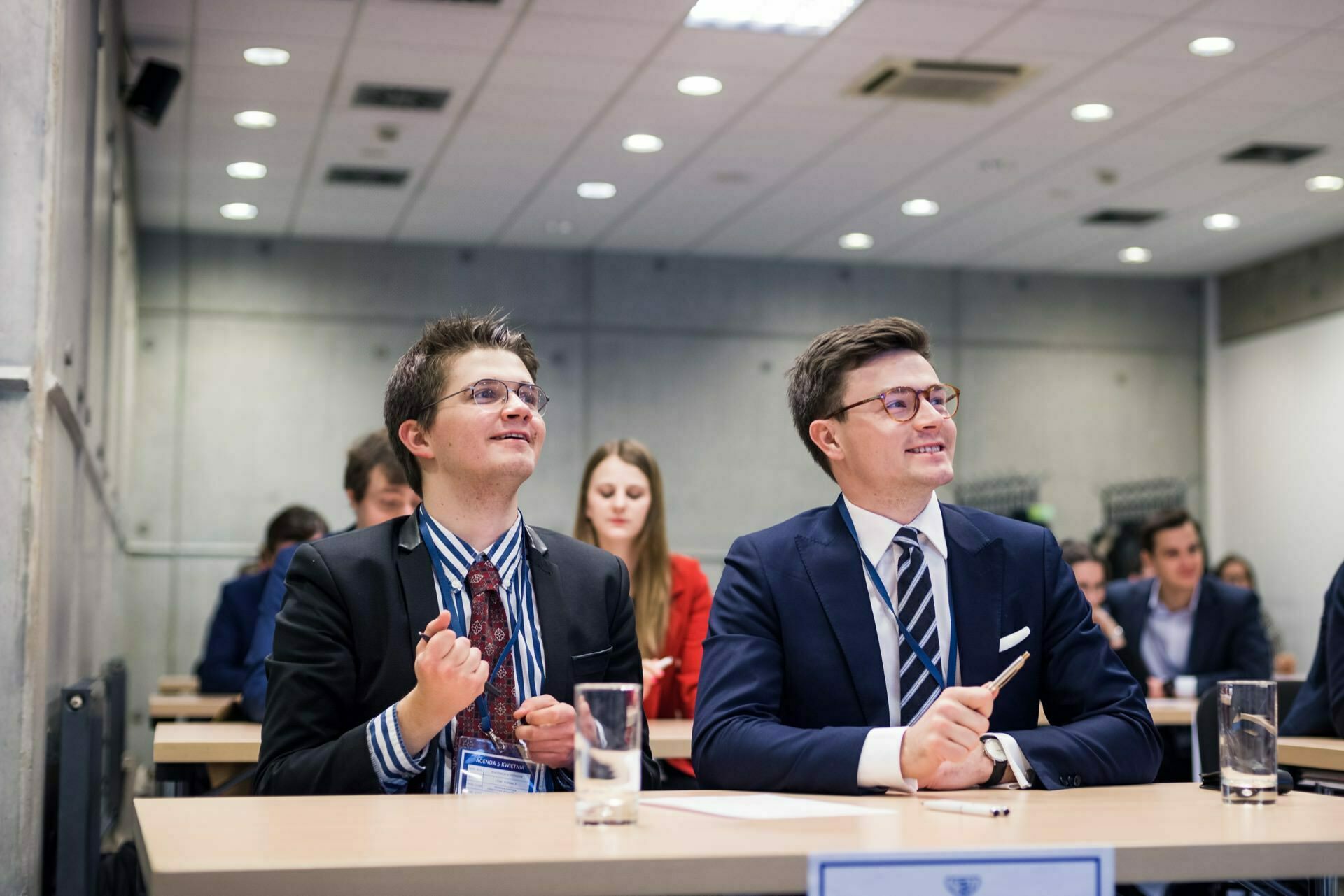 A group of people dressed in business attire sit at desks in a conference room. Two men in the foreground are smiling and look engaged, one is clenching his fists enthusiastically. The modern, professional setting enhances the photo coverage of the events with gray wall and ceiling lights.  