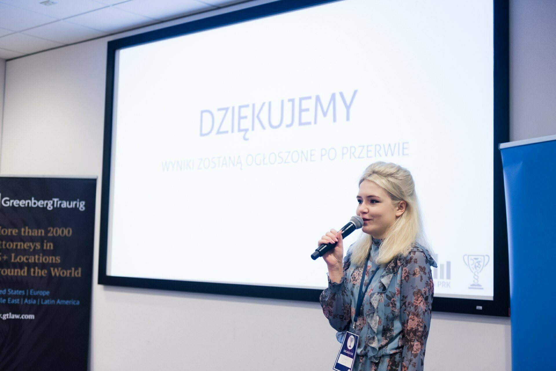 A woman stands in front of a screen with a microphone and makes a presentation. The screen displays the text "Thank you" and "Results properly announced when completed." On the left is a partially visible Greenberg Traurig banner, perfectly captured by an event photographer in Warsaw during an event photo shoot.  