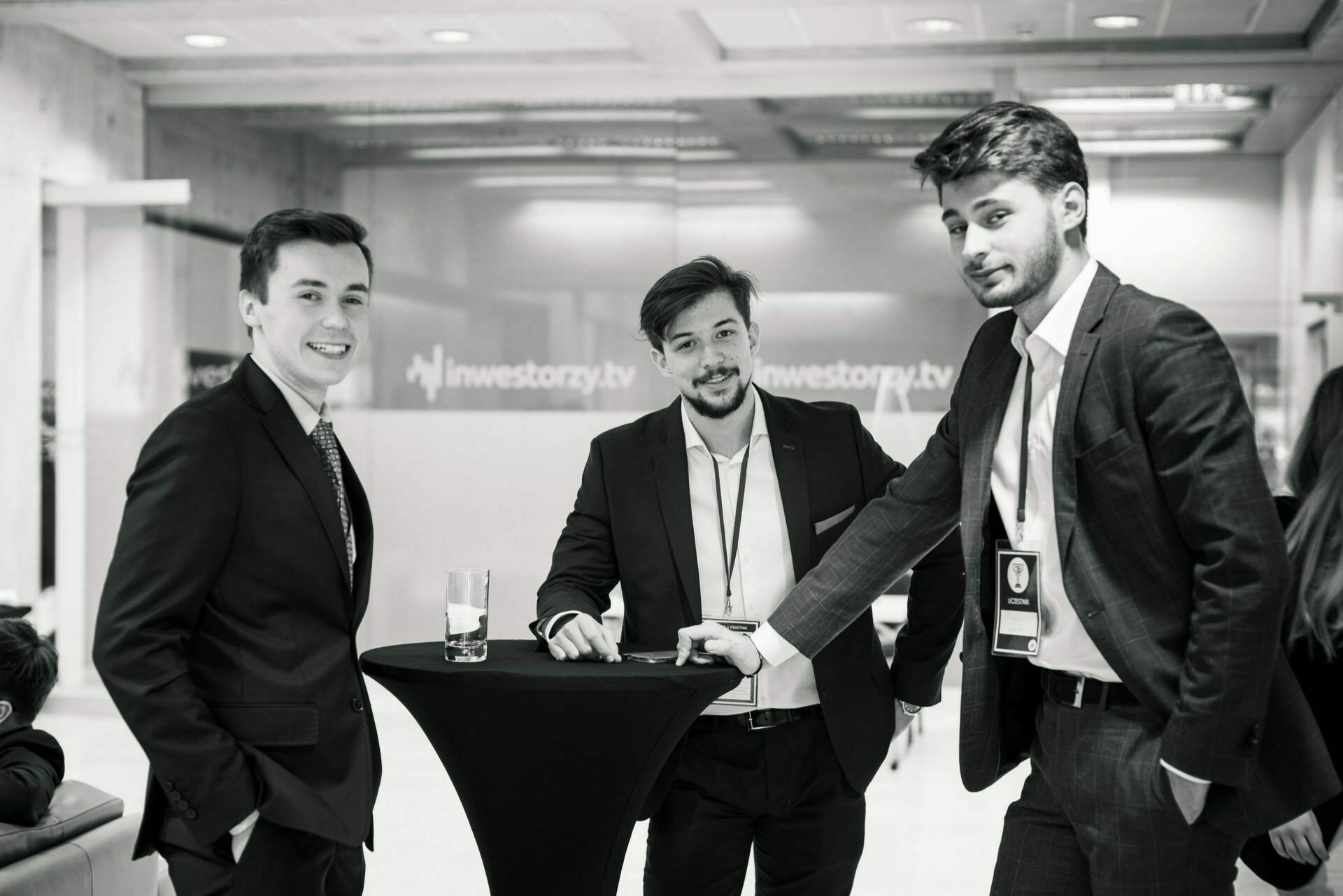 Black and white image of three men in formal attire standing around a small round cocktail table. They are smiling and appear busy in conversation. Two of the men are holding name badges, and there is a drink on the table. The background is a wall of frosted glass, which captures the essence of event photography.   