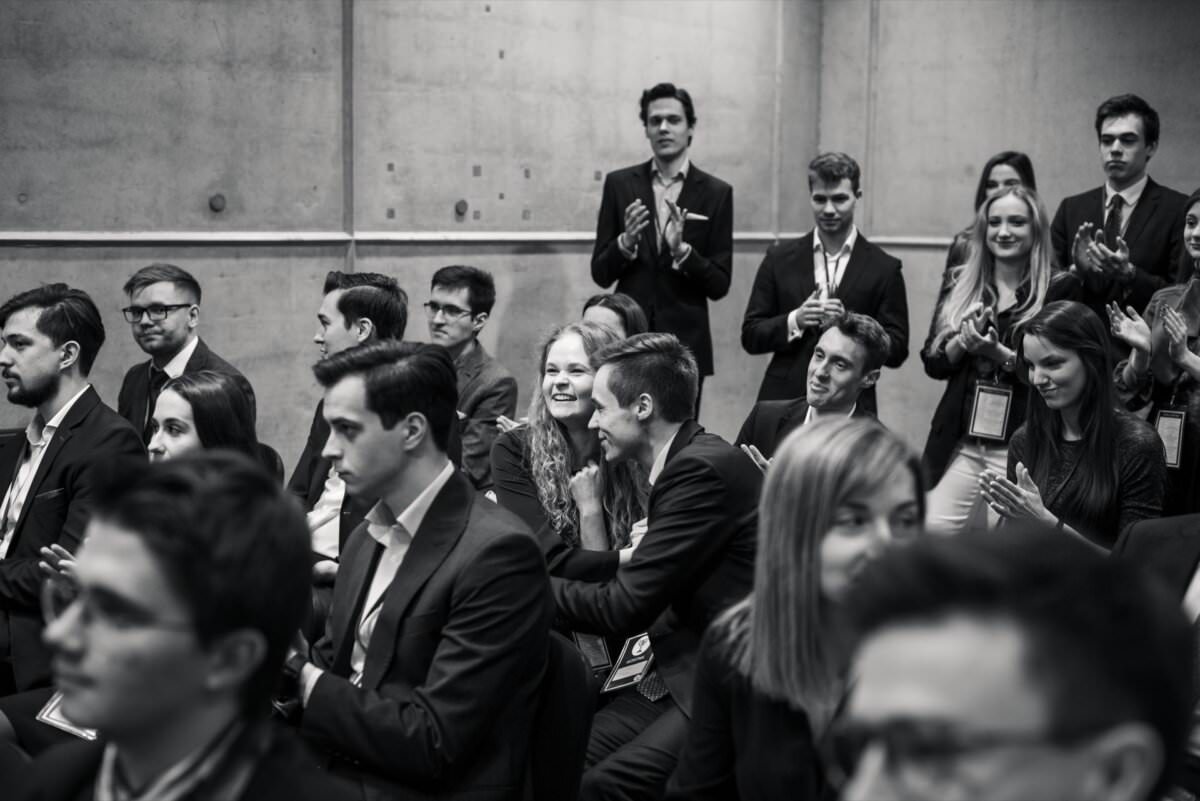 Schwarz-Weiß-Bild, das eine Gruppe formell gekleideter Menschen zeigt, die sitzen und stehen. Einige klatschen, andere unterhalten sich oder schauen sich aufmerksam an. Im Hintergrund ist eine Betonmauer zu sehen, die der Szene eine intime, professionelle Atmosphäre verleiht - das perfekte Eventfoto, aufgenommen vom Eventfotografen Warschau.