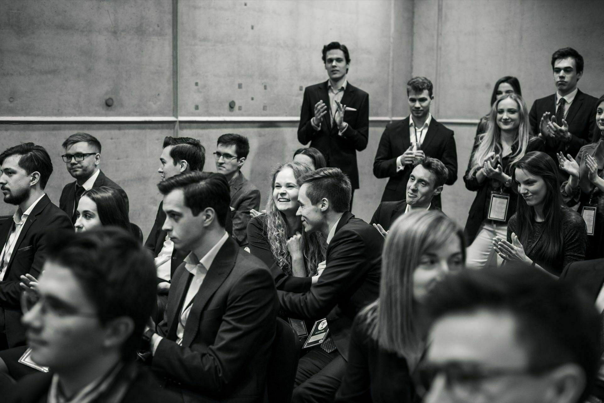 Black and white image showing a group of people dressed in formal attire, sitting and standing. Some are clapping, others are talking or watching intently. In the background is a concrete wall, which gives the scene an intimate, professional atmosphere - the perfect event photo captured by event photographer Warsaw.  