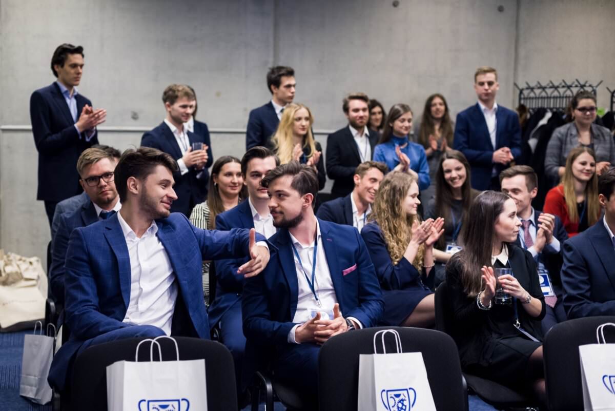 Eine Gruppe von Fachleuten in Anzügen sitzt und steht in einem Konferenzraum. Zwei Männer in der ersten Reihe schütteln sich die Hände und lächeln. Andere um sie herum klatschen, knüpfen Kontakte und schenken der Veranstaltung ihre Aufmerksamkeit. Auf den Stühlen sind Geschenktüten mit Logos platziert - ein perfektes Beispiel für Eventfotografie.