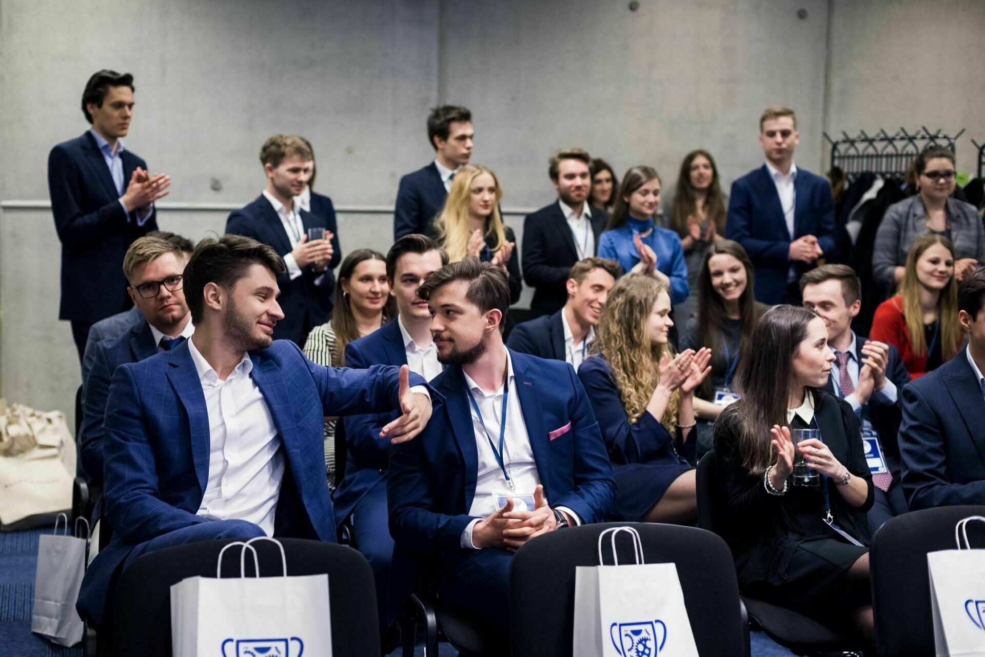 A group of professionals dressed in suits sit and stand in a conference room. Two men in the front row shake hands and smile. Others around them are clapping, socializing and paying attention to the event. Gift bags with logos are placed on the chairs - a perfect example of event photography.   