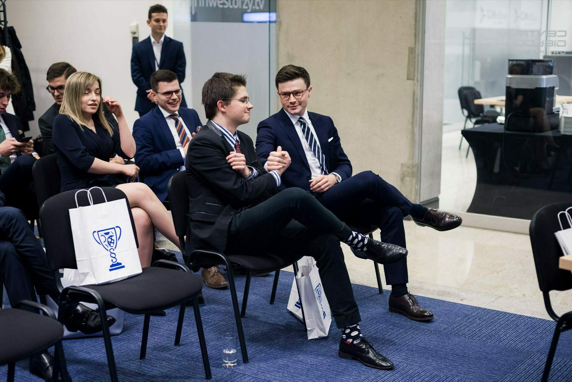 In a modern conference room sits a group of people dressed in formal attire. In the foreground sit two men in suits, immersed in conversation. Several branded gift bags are set on the floor next to the chairs, creating the perfect setting for event photo coverage by any event photographer Warsaw has to offer.  