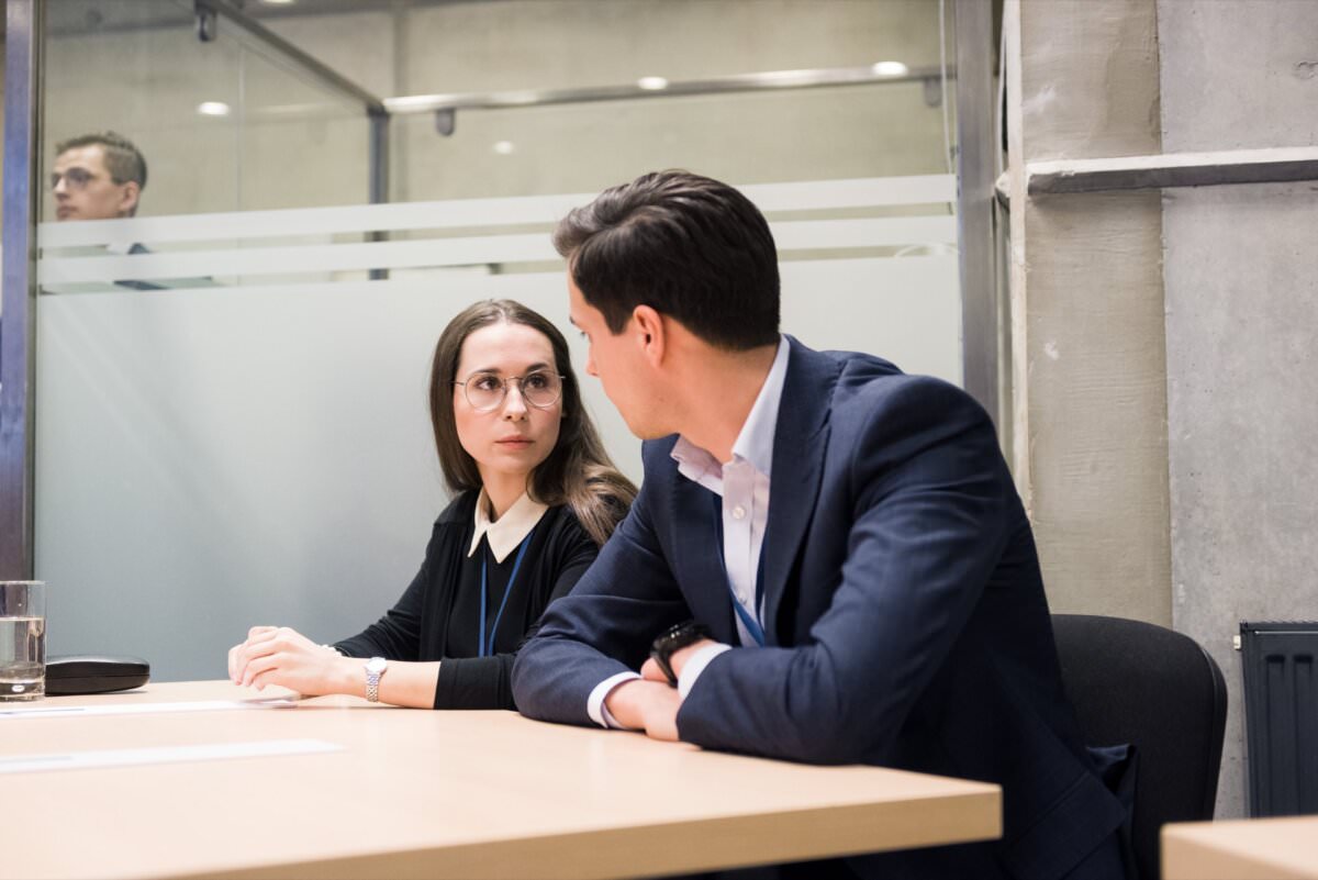 Zwei Berufstätige sitzen an einem Schreibtisch in einem Bürokonferenzraum und unterhalten sich. Eine Frau mit Brille und schwarzem Pullover schaut einen Mann in einem marineblauen Anzug an. Ein Glas Wasser und Dokumente liegen auf dem Tisch. Der Raum, den der Eventfotograf warszawa perfekt eingefangen hat, ist mit Glastrennwänden ausgestattet.